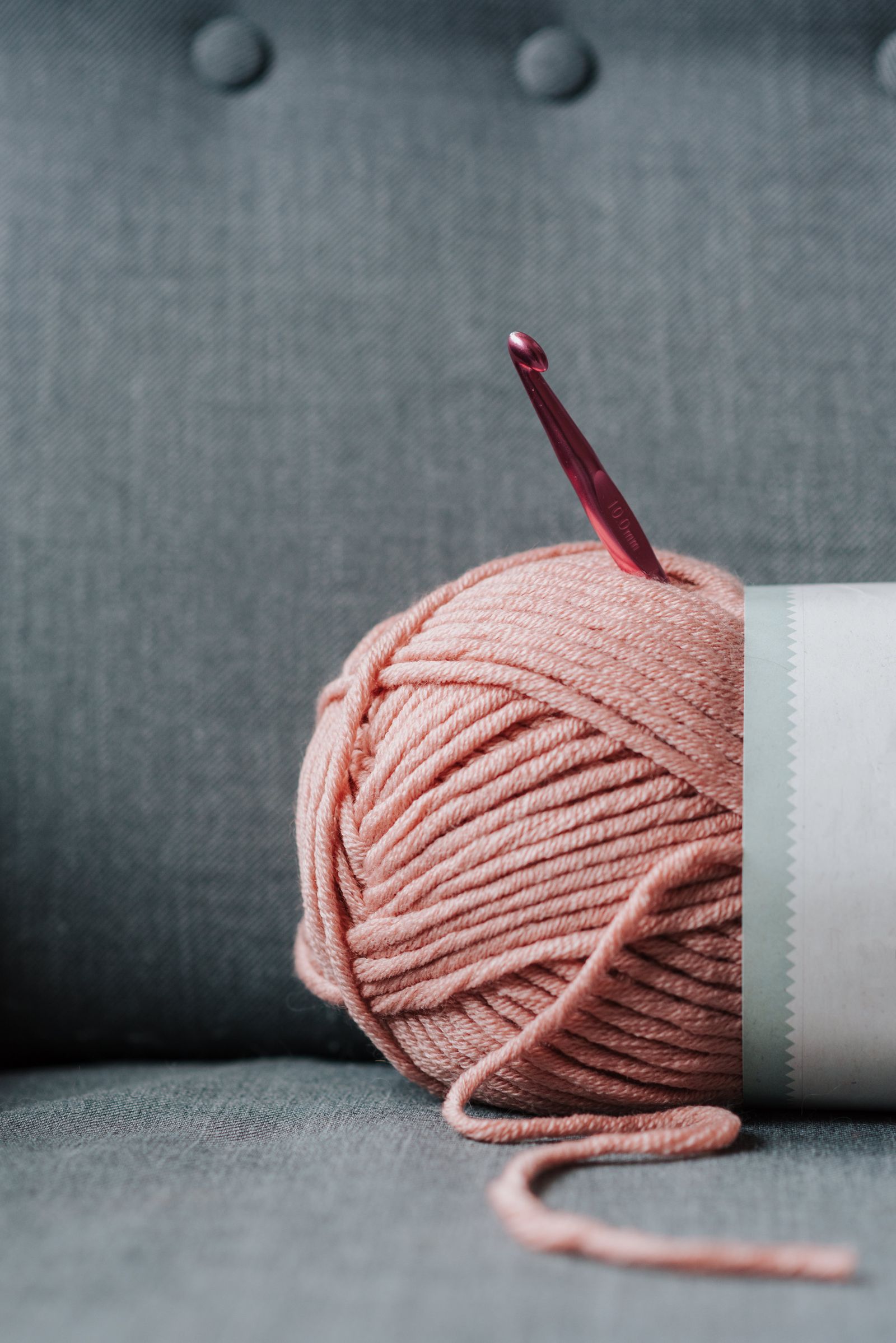 Ball of soft yarn with pink crochet hook on a cozy sofa